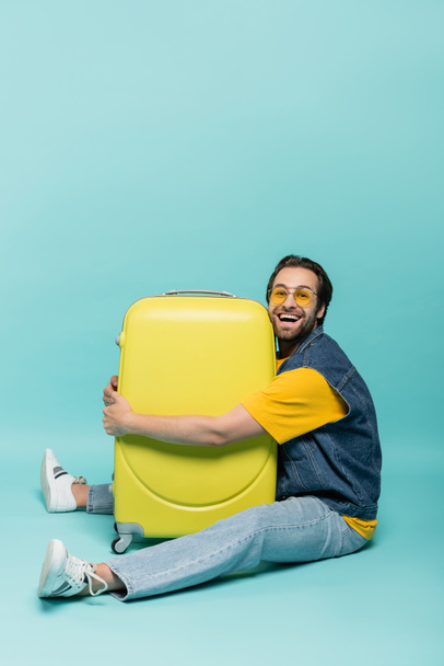 Cheerful man hugging suitcase on blue background  - Photo, Image