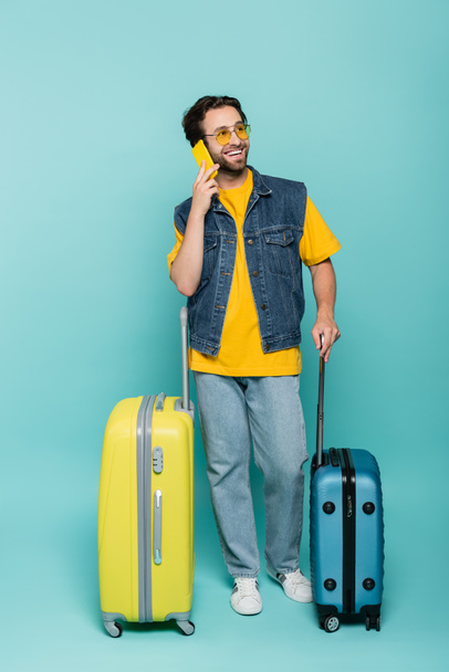 Smiling man in sunglasses talking on smartphone near suitcases on blue background  - Photo, image