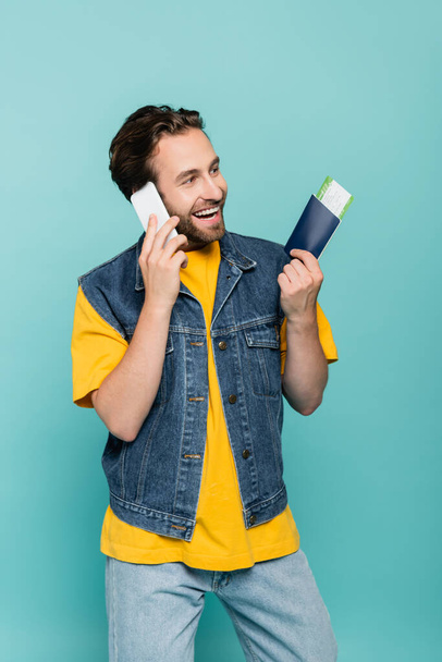 Cheerful tourist with passport talking on smartphone isolated on blue  - Foto, imagen