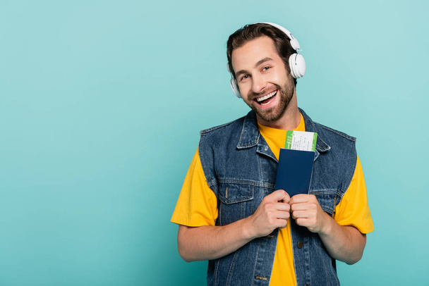 Χαμογελώντας ταξιδιώτη με ακουστικά που κατέχουν διαβατήριο με αεροπορικό εισιτήριο απομονώνονται σε μπλε  - Φωτογραφία, εικόνα