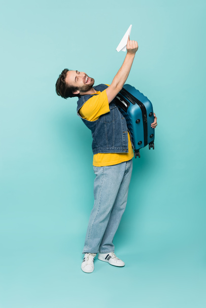 Laughing man holding suitcase and paper plane on blue background  - Foto, immagini