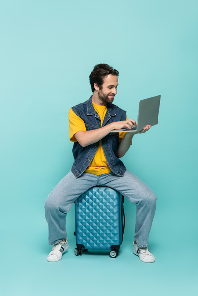 Smiling freelancer using laptop on suitcase on blue background  - Foto, immagini