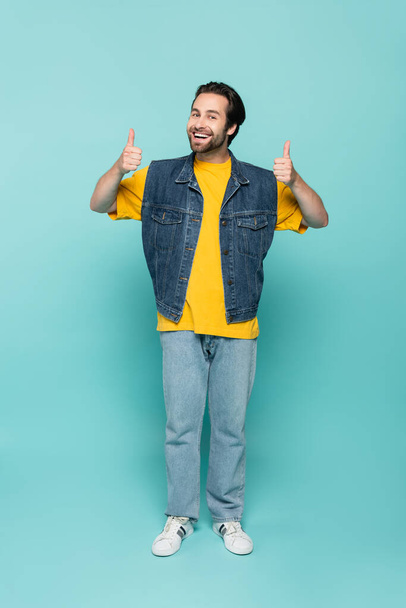 Smiling man in denim vest showing thumbs up on blue background  - Fotoğraf, Görsel