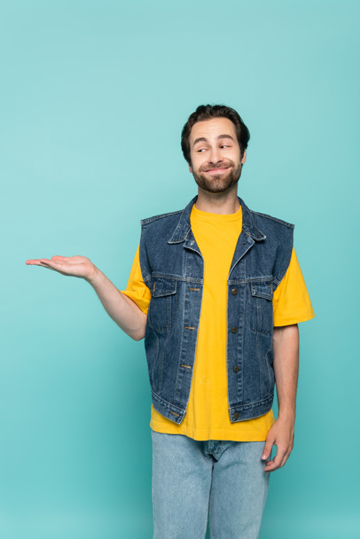Smiling man in t-shirt and denim vest pointing with hand isolated on blue  - 写真・画像