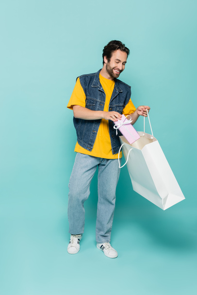 Young man putting present in shopping bag on blue background  - Фото, зображення