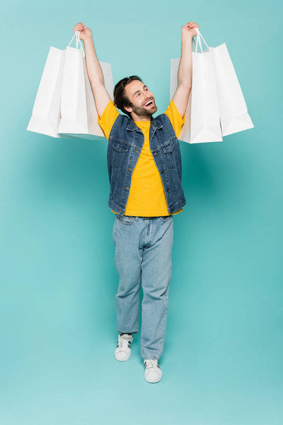Full length of smiling man rising shopping bags on blue background  - 写真・画像