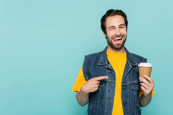 Giovane uomo sporgente lingua mentre indica il caffè per andare isolato su blu - Foto, immagini