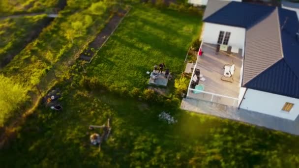 Tiro de dron de ángulo alto de una casa en una pequeña ciudad con patio verde donde el hombre está haciendo barbacoa - Metraje, vídeo