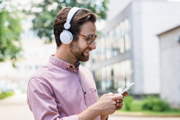 Side view of smiling businessman in shirt using smartphone and headphones outdoors  - Valokuva, kuva