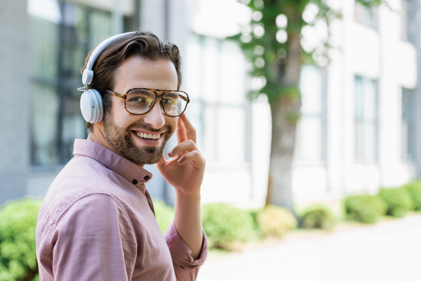 Smiling businessman in eyeglasses and headphones looking at camera outdoors  - Photo, Image