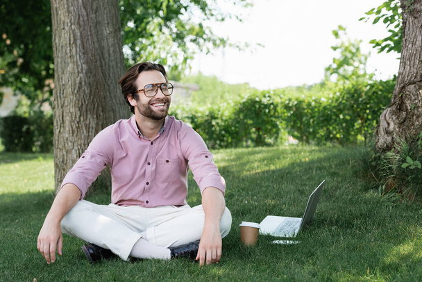 Happy businessman sitting near devices and paper cup on grass  - Фото, зображення