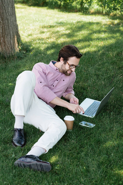 High angle view of smiling businessman using laptop near takeaway drink and smartphone on grass  - Photo, Image