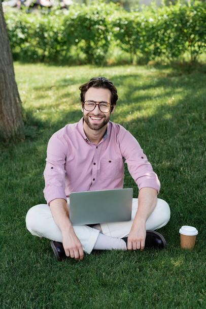 Uomo d'affari positivo con computer portatile guardando la fotocamera vicino tazza di carta su erba  - Foto, immagini