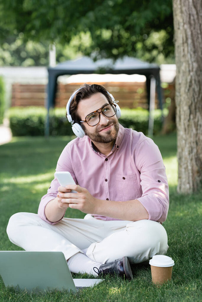Dreamy businessman in headphones using gadgets near paper cup on grass  - Фото, изображение