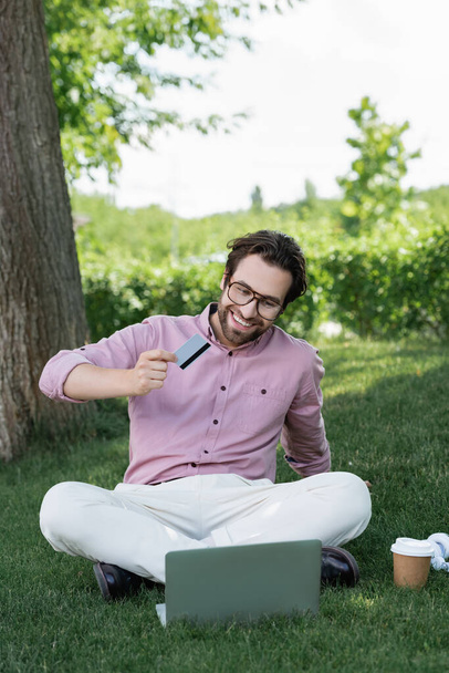 Veselý podnikatel držící kreditní kartu během videohovoru na notebooku na trávě  - Fotografie, Obrázek