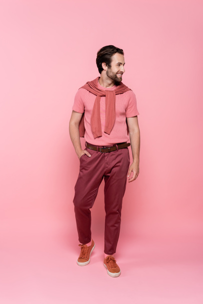 Pleine longueur d'homme souriant tenant la main dans une poche de pantalon sur fond rose - Photo, image
