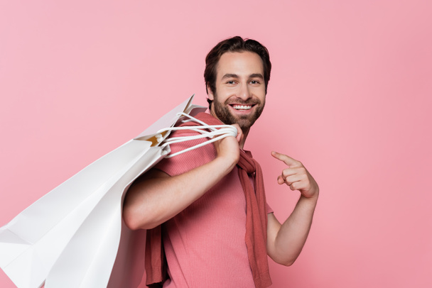 Pozitivní muž ukazuje na nákupní tašky izolované na růžové - Fotografie, Obrázek