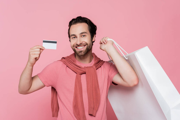 Young man showing credit card while holding shopping bag isolated on pink - Foto, Imagem