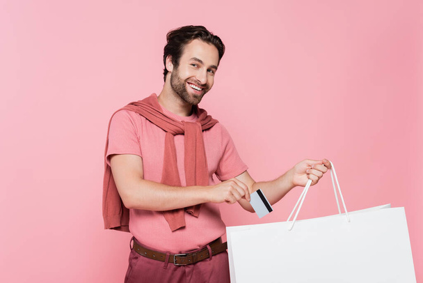 Smiling man putting credit card in shopping bag isolated on pink - Foto, afbeelding