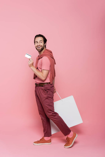 Full length of shopaholic with credit card and shopping bag walking on pink background - Foto, afbeelding