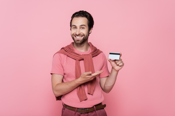 Hombre positivo apuntando a la tarjeta de crédito mientras está de pie aislado en rosa - Foto, Imagen