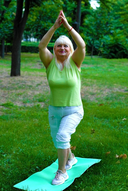 Starší blondýnka cvičí jógu v parku na rohožce. Starší blondýnka chodí na sport do parku. Starší žena dělá fyzickou aktivitu v parku a vede zdravý životní styl.  - Fotografie, Obrázek
