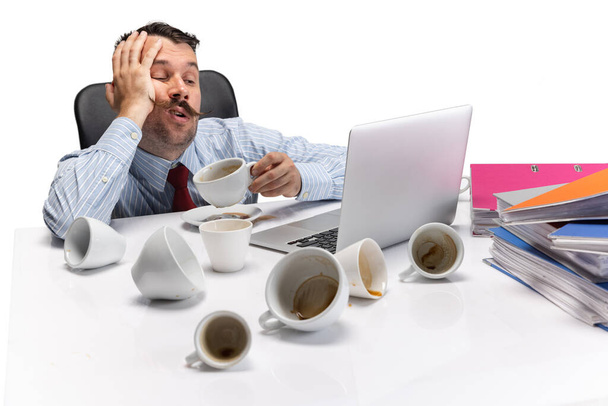 Retrato de jovem homem caucasiano sonolento e cansado, trabalhador de escritório sentado à mesa sobre fundo branco. - Foto, Imagem