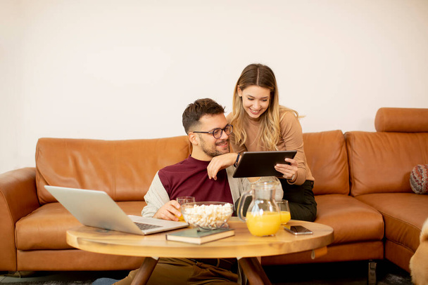 Schönes junges Paar benutzt Laptop zusammen, während es zu Hause auf dem Sofa sitzt - Foto, Bild