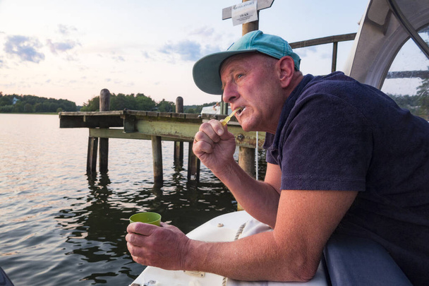 Een man van middelbare leeftijd poetst zijn tanden op het achterste gedeelte van een kleine motorboot tijdens het kamperen. - Foto, afbeelding