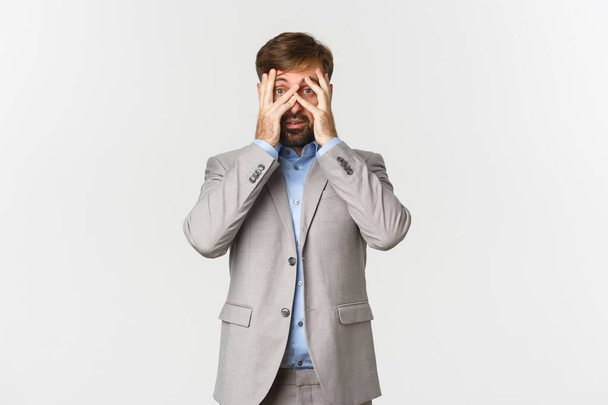 Portrait of ambushed businessman in grey suit, cover eyes and peeking through fingers with scared face, standing over white background - Photo, Image