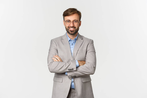 Image of successful and confident businessman with beard, wearing grey suit and glasses, cross arms on chest and smiling satisfied, standing over white background - Photo, Image