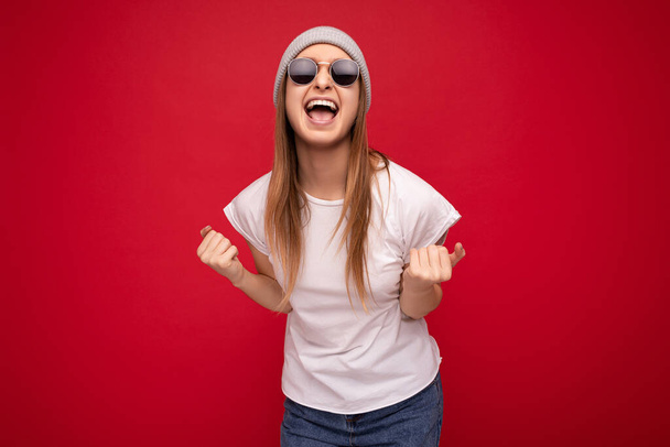 Photo shot of attractive happy joyful young blonde female person isolated over red background wearing casual white t-shirt for mockup and stylish sunglasses looking at camera and celebrating winning - Photo, Image