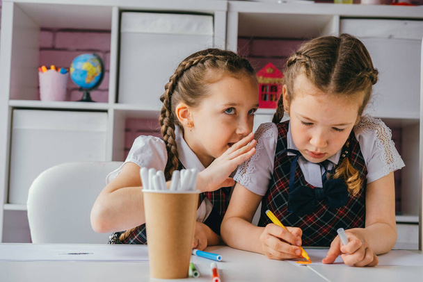 Two girls are sitting at a desk in the classroom at the lesson. One girl whispers gossip to another girl. School friendship concept. - Фото, зображення