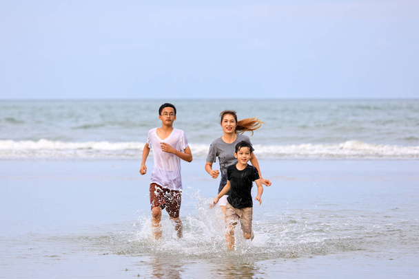Arka planda mavi gökyüzü olan tropikal plajda birlikte koşan üç Asyalı aile. - Fotoğraf, Görsel