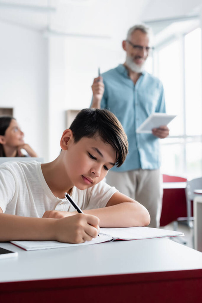 Asian schoolboy writing on notebook near blurred teacher  - Фото, изображение