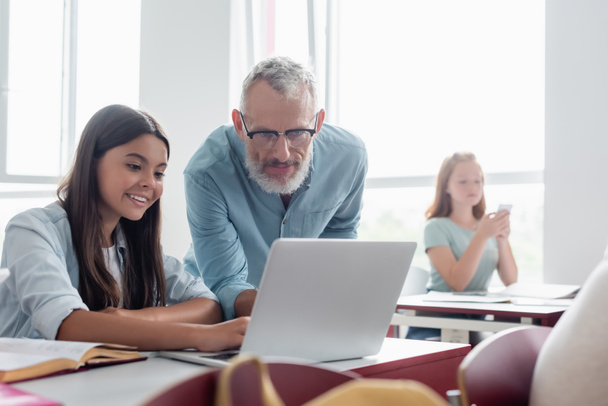 Teacher looking at laptop near smiling schoolgirl in classroom  - Foto, Imagen