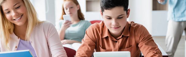 Schulkind blickt auf verschwommenes digitales Tablet neben lächelndem Klassenkameraden, Banner  - Foto, Bild