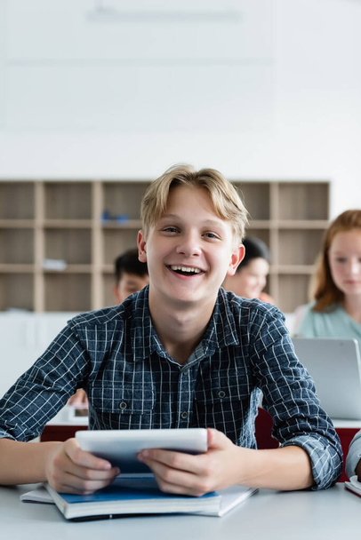 Happy schoolboy holding digital tablet and looking at camera in classroom  - Foto, afbeelding