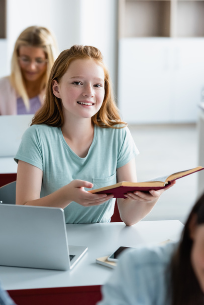 Smiling schoolgirl holding book near devices in classroom  - Photo, Image