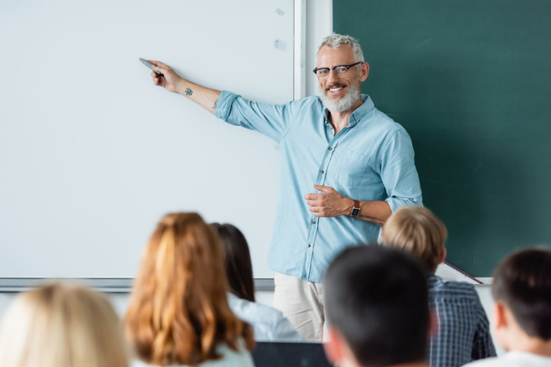Smiling teacher pointing at erase board near blurred pupils  - 写真・画像
