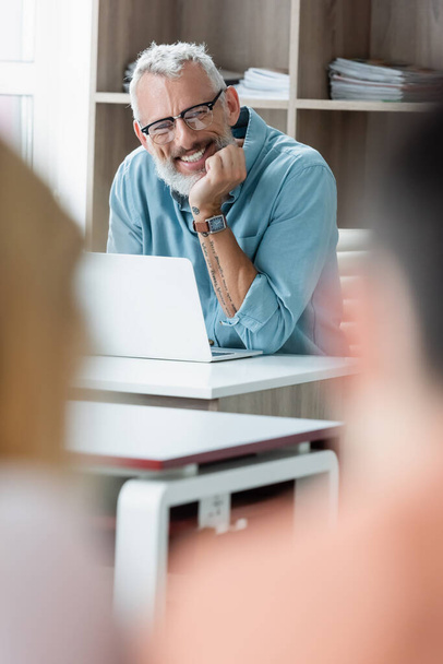 Sonriente profesor maduro sentado cerca de la computadora portátil en la escuela  - Foto, Imagen