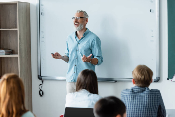 Profesora positiva con marcador hablando cerca del tablero de borrado y escolares en el aula  - Foto, imagen