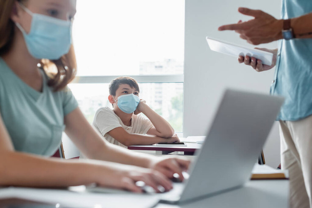 Asian schoolkids in medical mask looking at blurred teacher with digital tablet  - Valokuva, kuva