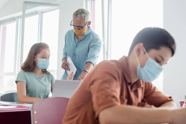 Mature teacher in medical mask standing near schoolgirl pointing at laptop during lesson  - Photo, Image