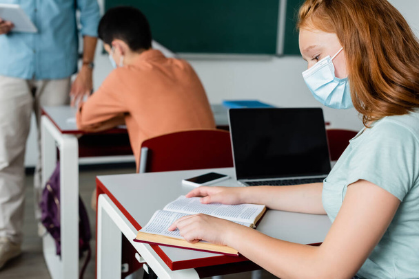 Schoolgirl in medical mask reading book near devices in classroom  - Fotó, kép