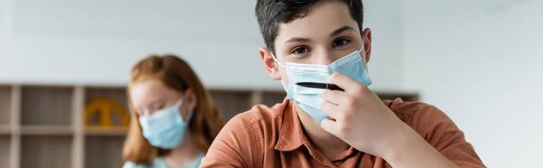Écolier en masque médical tenant un stylo dans la salle de classe, bannière  - Photo, image