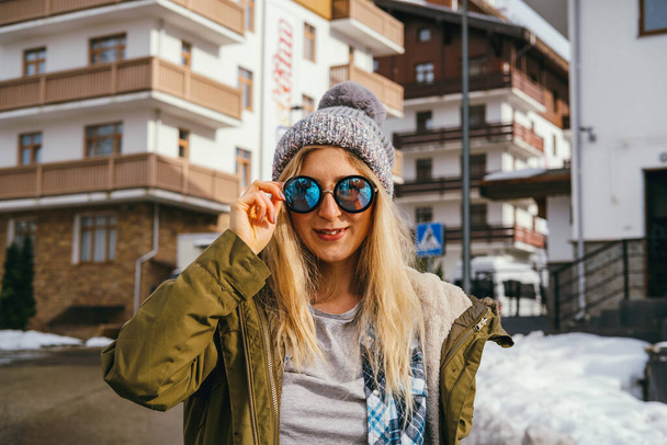 Młoda szczęśliwa kobieta nosi okulary przeciwsłoneczne, pom pom hat i kurtkę. Fajny hipster kobieta portret na zewnątrz - Zdjęcie, obraz