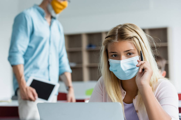 Schoolgirl in medical mask holding pen near laptop and blurred teacher  - Fotó, kép