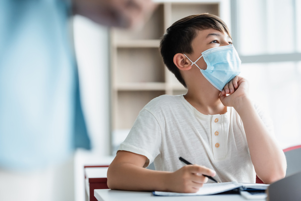 Aziatisch schooljongen in medisch masker houden pen in de buurt wazig leraar  - Foto, afbeelding