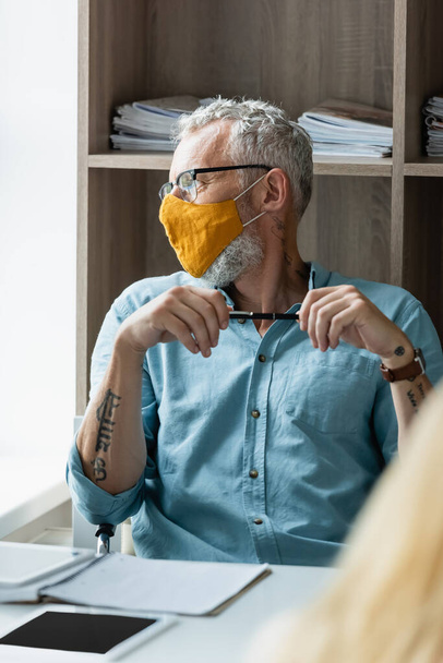 Teacher in protective mask holding pen near notebooks and digital tablets on table  - Fotoğraf, Görsel